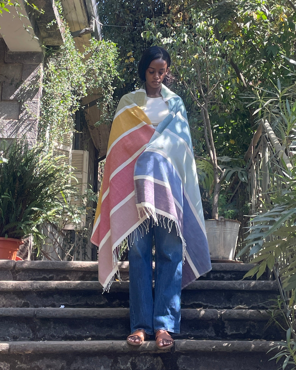 A woman stands on outdoor stone steps surrounded by lush green plants and trees, wrapped in a striped multicolor blanket with white fringe. She is wearing blue jeans and sandals.