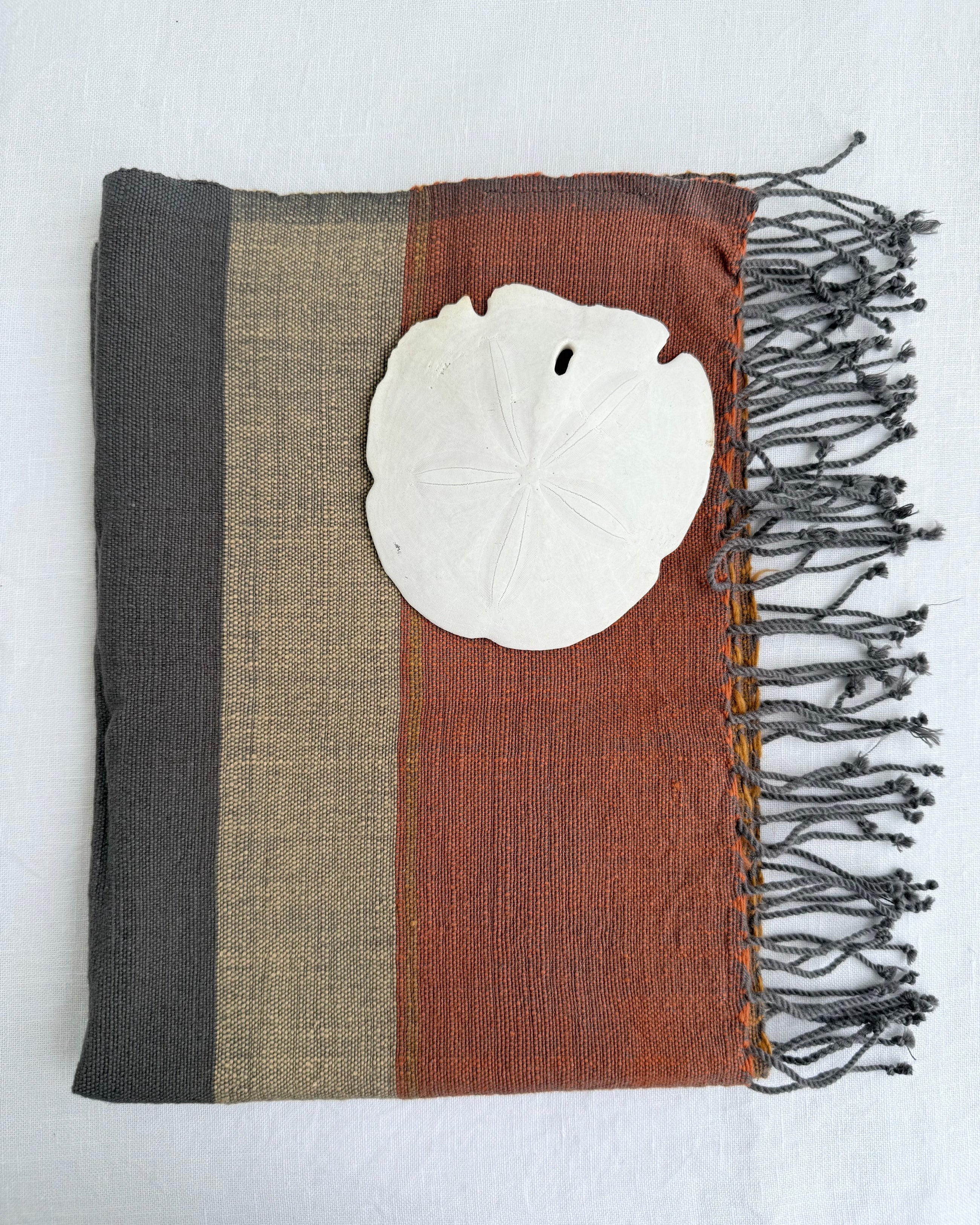 A square woven hand towel with vertical stripes in dark gray, beige, and rust brown, finished with dark fringe on one side and a white shell placed on top, shown against a white background.