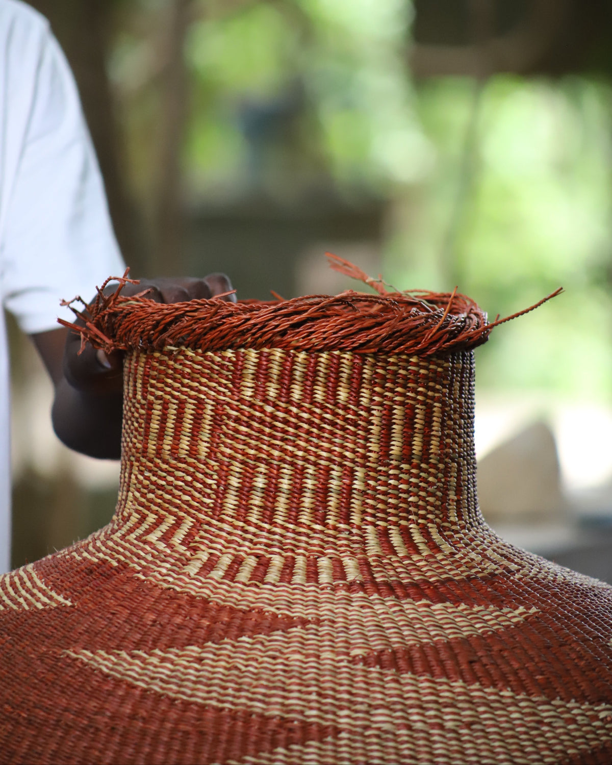 A close-up of a hand-woven basket with a wide, rounded body and a narrow neck, decorated with geometric patterns in reddish-brown and beige. A person's arm and white shirt are visible on the left side, with a blurred outdoor background of green foliage.