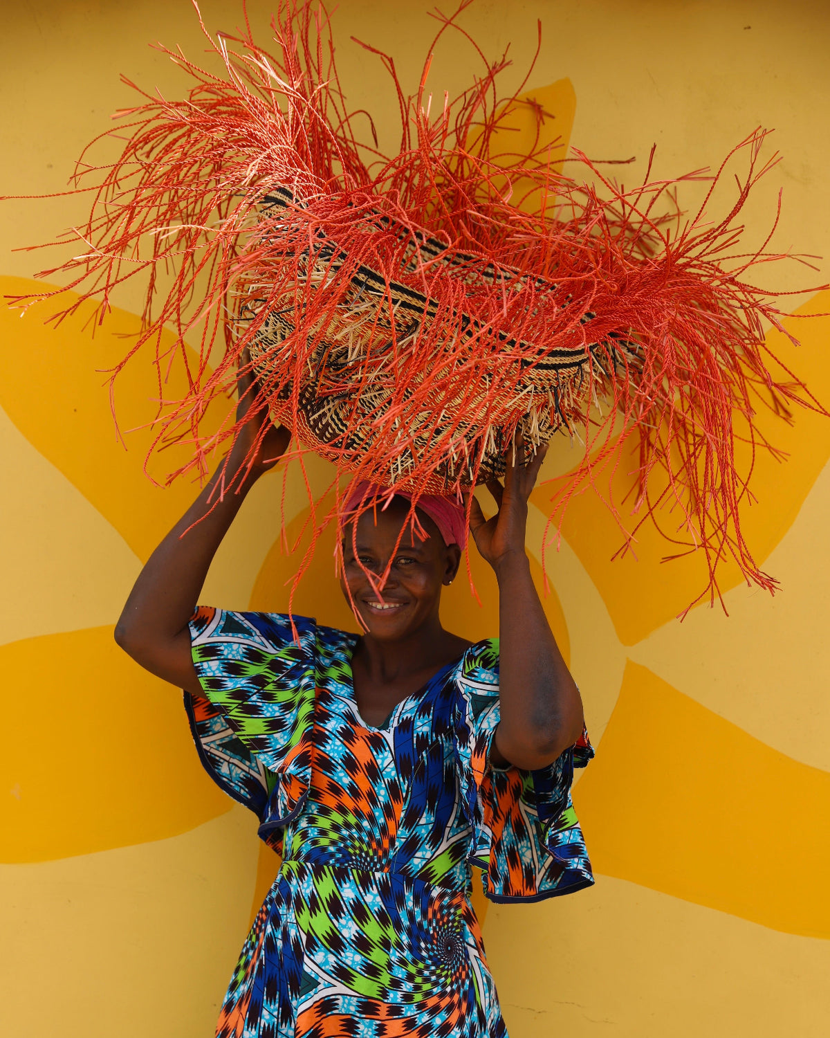 A person holding a colorful basket as headwear with a vibrant red and yellow design, holding the hat up against a yellow background.