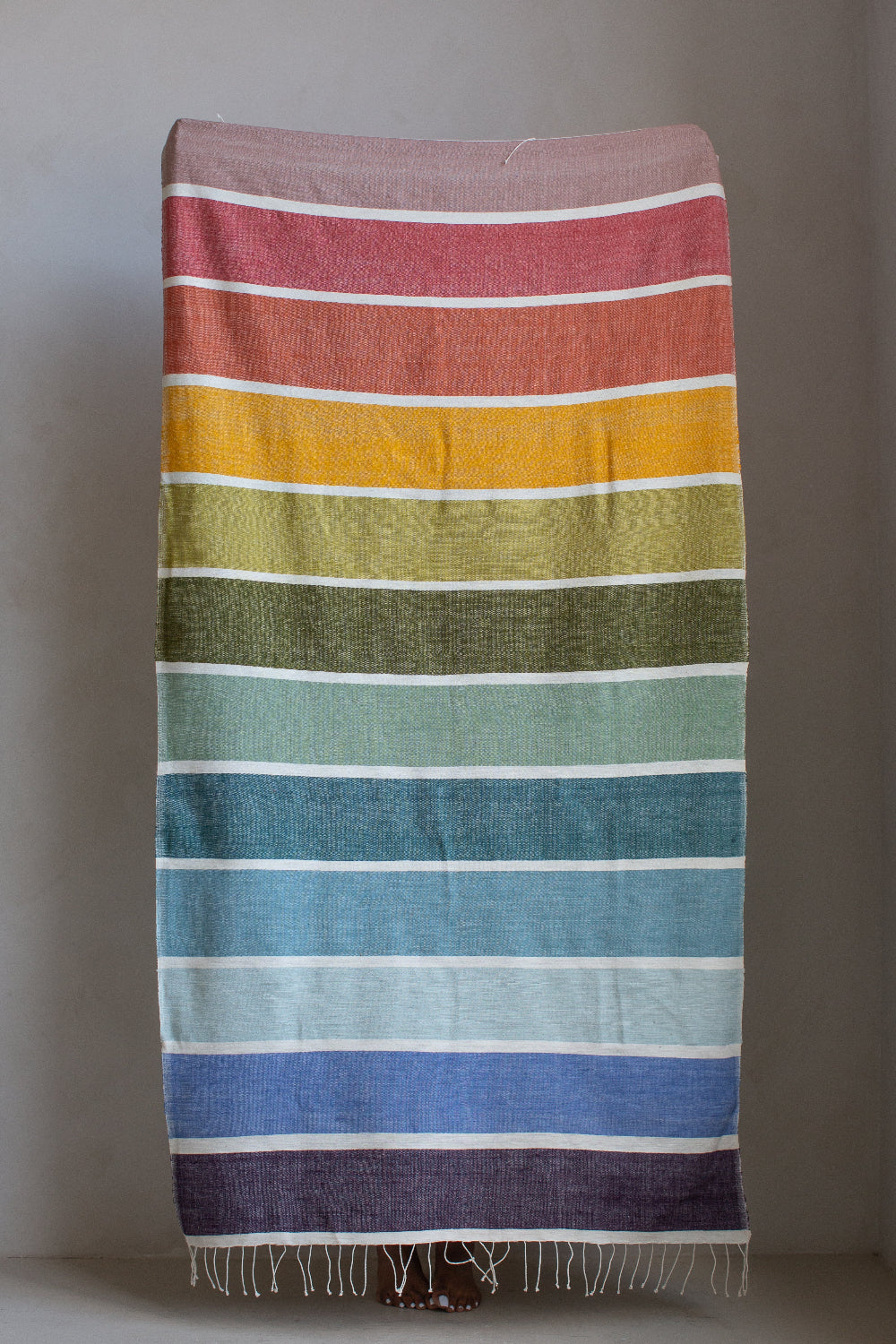 A rectangular woven textile with horizontal rainbow stripes in pink, orange, yellow, olive green, teal, blue, and purple, hanging against a plain gray wall with fringe along the bottom edge.