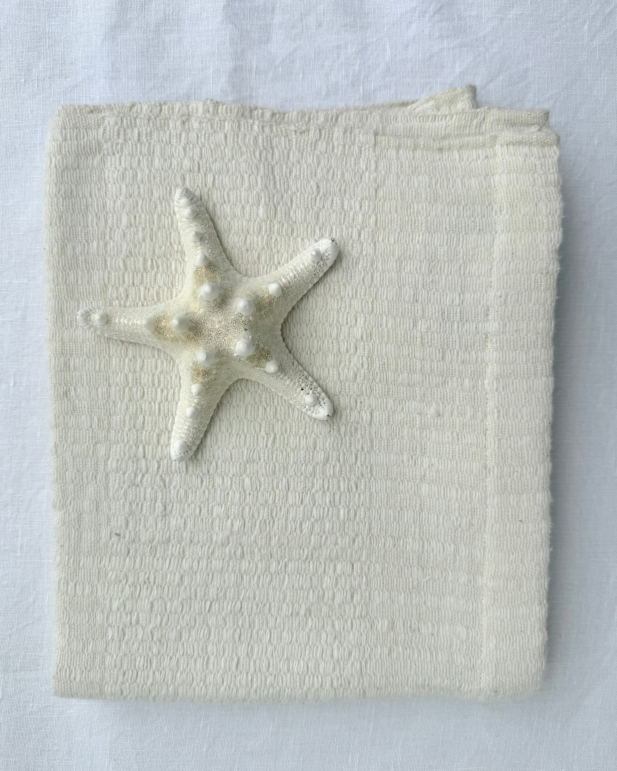 A folded cream-colored hand towel, featuring a small white starfish decoration on the front, set against a matching white fabric background.