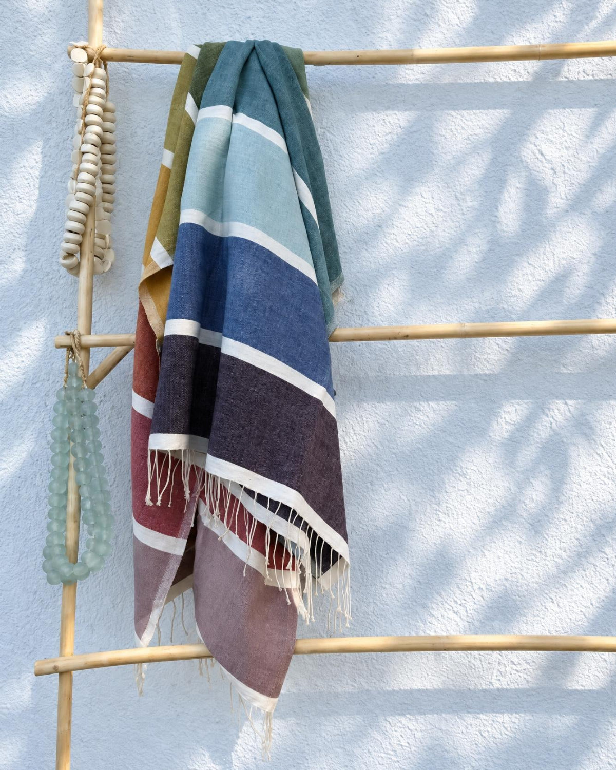 A striped woven throw blanket with fringe, draped over a bamboo rack against a white textured wall with leaf shadows.