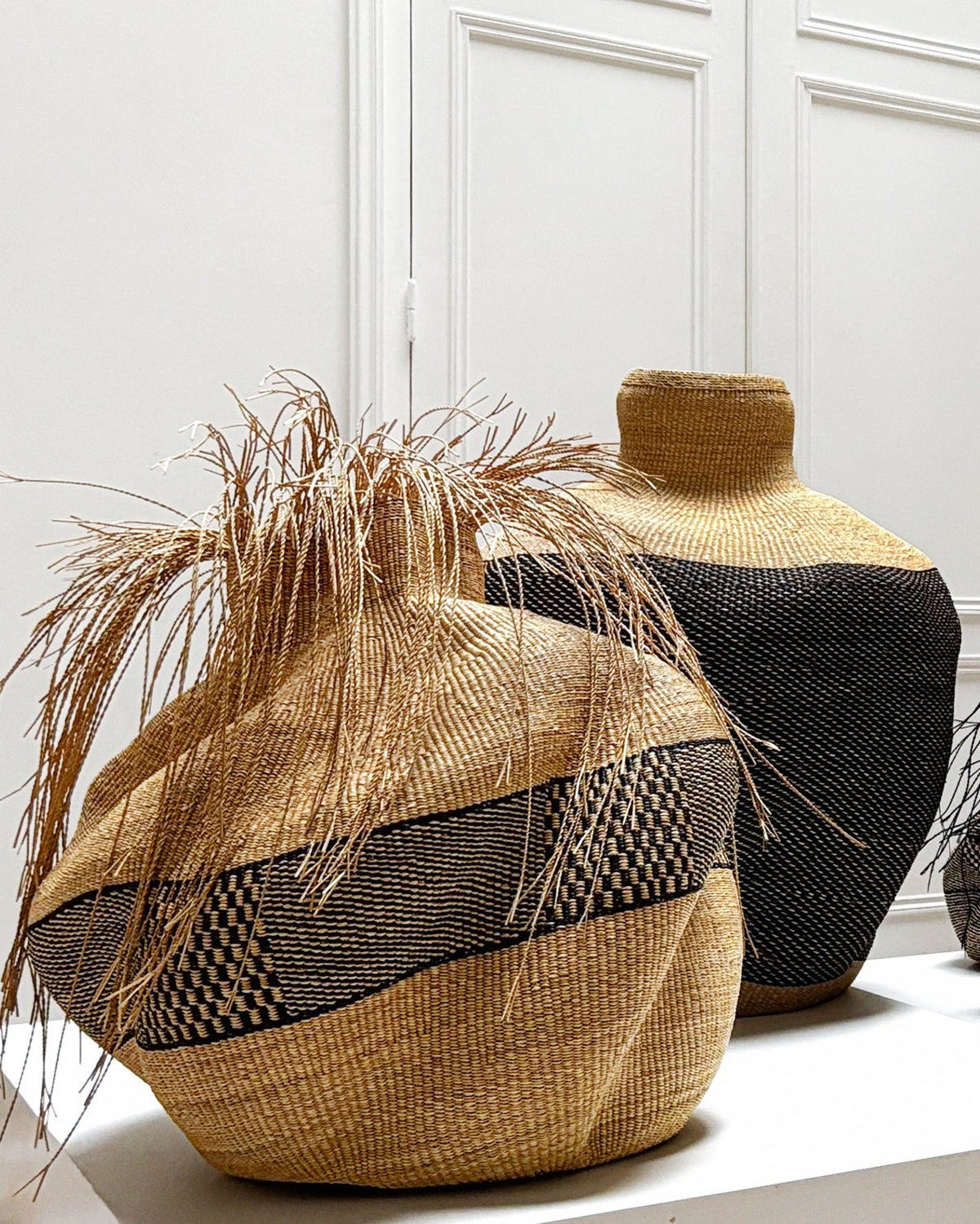 A collection of hand-woven art baskets vases and decorative pots displayed on a white gallery shelf against a white paneled wall, with a framed Samburu portrait hanging in the background.