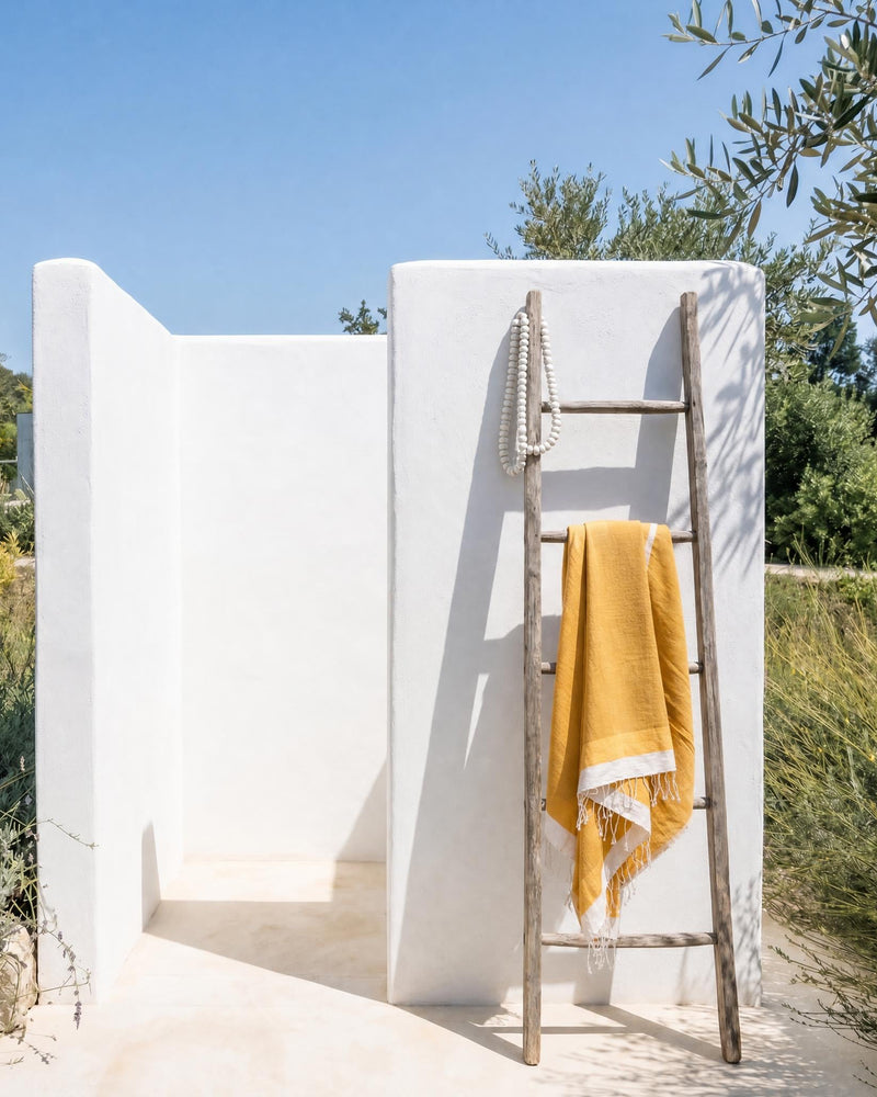 A minimalist outdoor shower with a yellow handwoven towel draped over a wooden ladder. The scene is set in a sunny, natural environment with green plants and a clear blue sky.