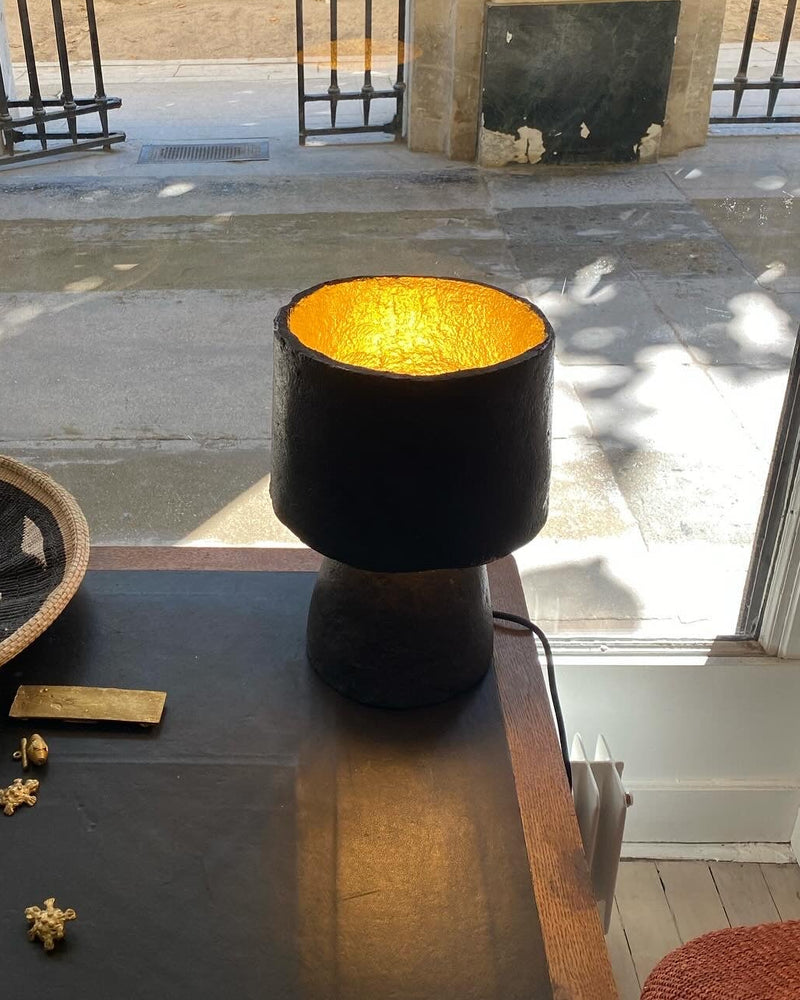 A black paper pulp table lamp with a glowing gold interior sits on a dark tabletop in a real-world setting, with a woven tray, jewelry, and an outdoor garden visible through a stone doorway in the background.