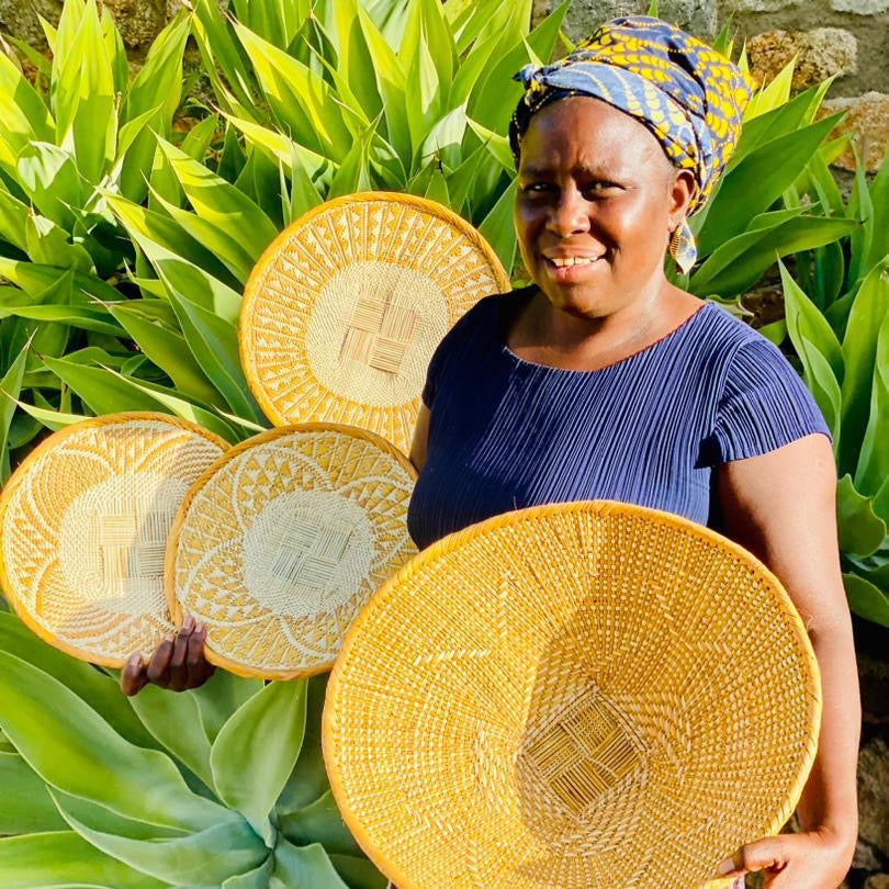 A smiling woman wearing a colorful headwrap and blue top holds four round woven baskets with intricate geometric patterns, set against a background of green succulents.