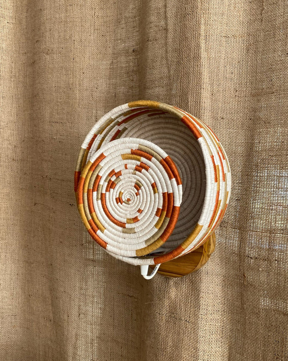 A hand-woven spiral basket with white, orange, and beige bands, displayed on a wooden stand against a textured burlap backdrop.