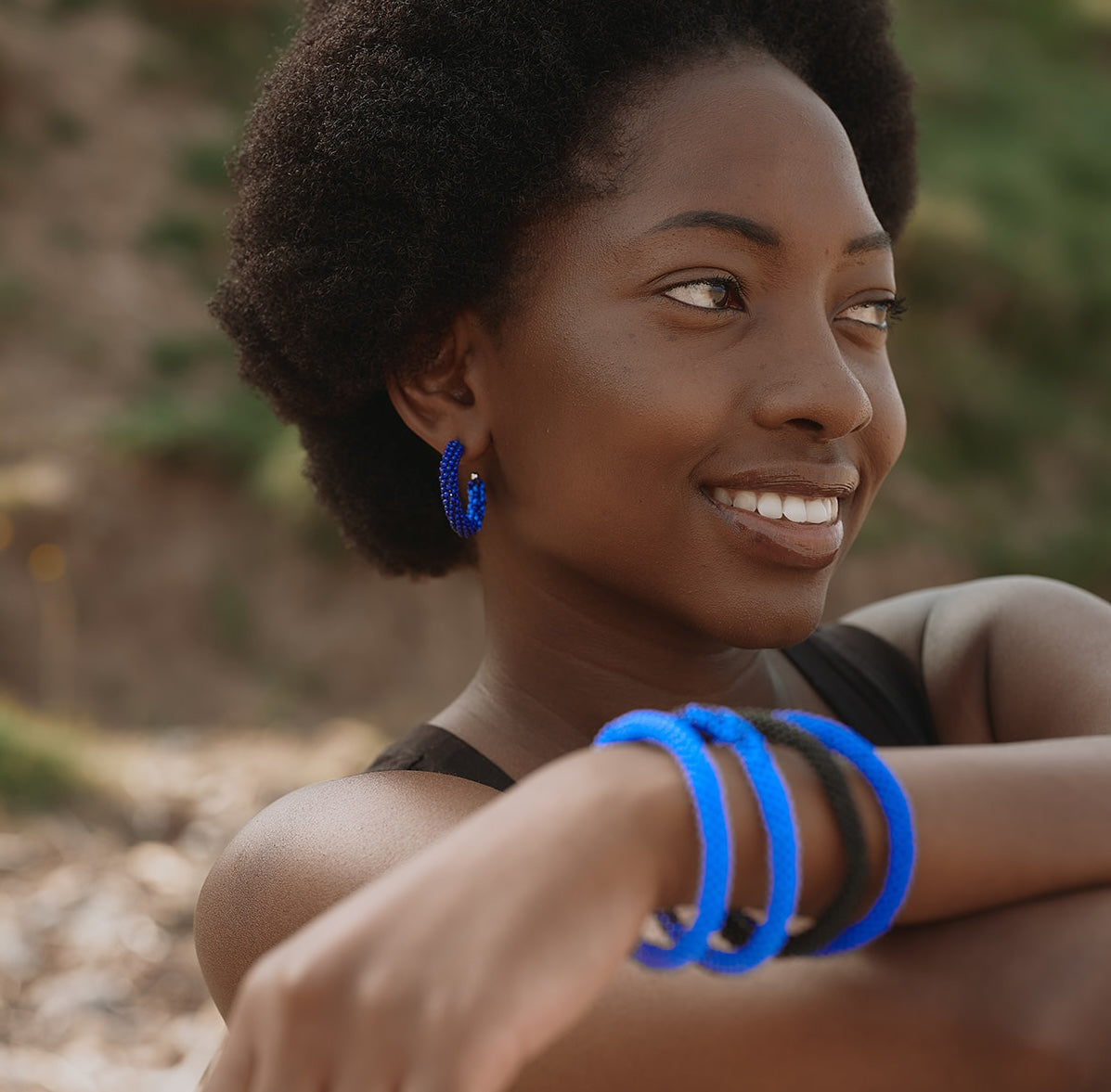 A woman outdoors wearing bright blue beaded hoop earrings and matching thick blue bangles on her wrist, with a blurred natural background.
