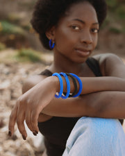 A woman wearing three braided blue and black bangles on her wrist, sitting outdoors with a blurred natural background.