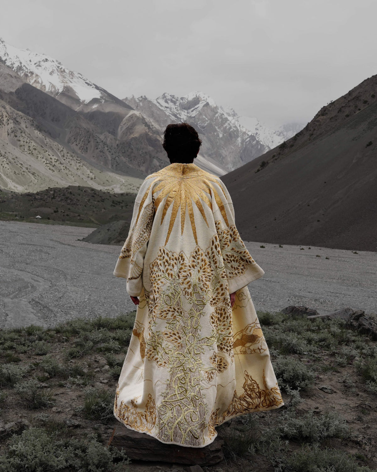 A person stands with their back to the camera in a vast mountain valley, wearing a long cream and gold patterned robe or cloak. Snow-capped mountains and a rocky riverbed stretch out in the background under an overcast sky.