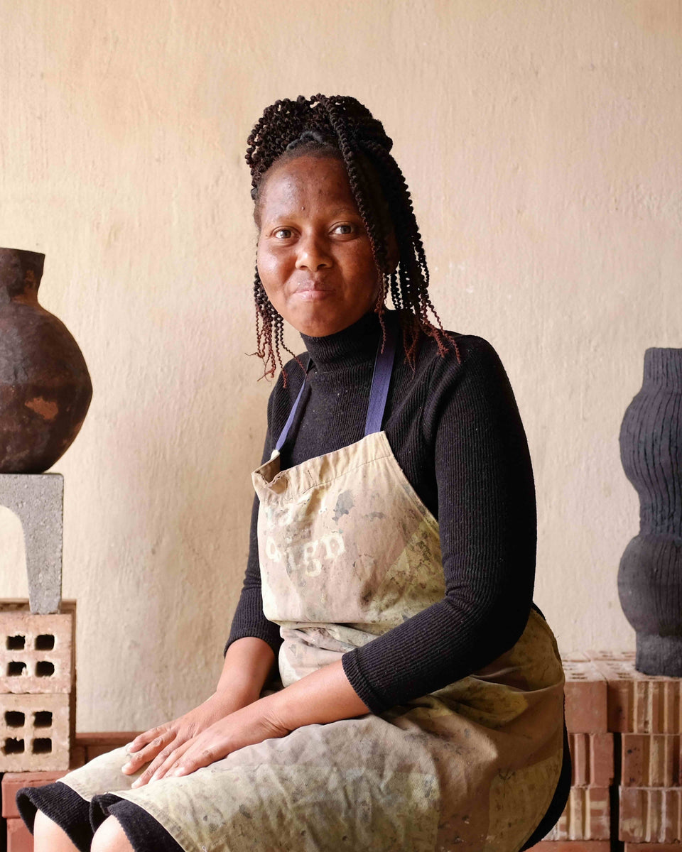 A woman wearing a beige apron over a black long-sleeve shirt sits in a pottery studio surrounded by handmade ceramic vases and sculptural vessels displayed on brick and concrete blocks against a beige plaster wall.
