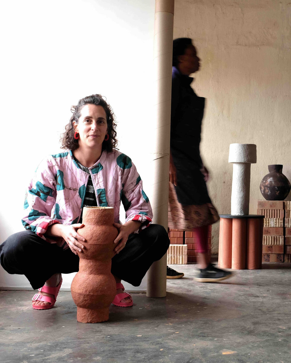 A woman crouches in a gallery space holding a terracotta-colored sculptural vase. The room features minimalist columns, a concrete floor, and other decorative ceramic vases displayed on pedestals in the background and to the right.