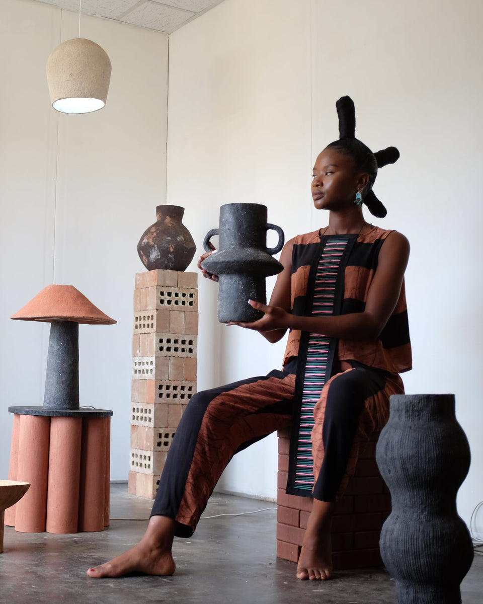 A woman in a patterned brown and black dress sits barefoot on a brick stool in a gallery space, holding a dark ceramic vessel. The room features various sculptural art pieces, terracotta tables, and a large wooden panel on the wall.