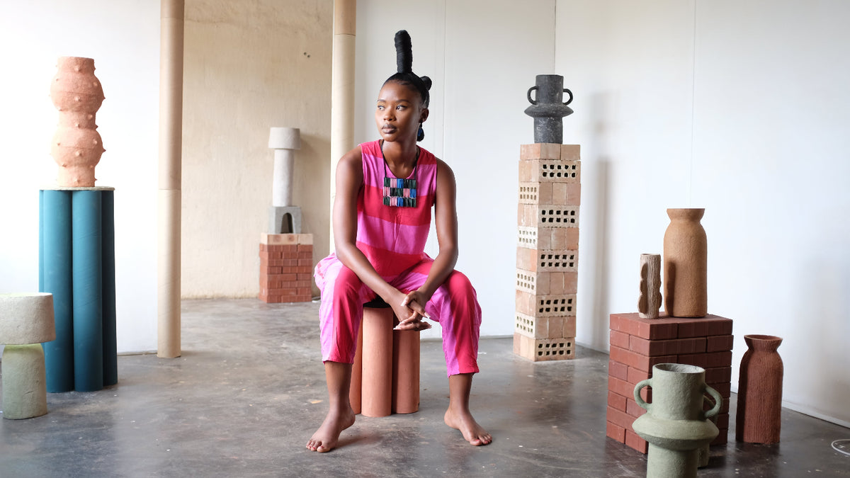 A model wearing a vibrant pink sleeveless jumpsuit with a colorful geometric chest pattern sits on a terracotta stool in a gallery space surrounded by sculptural ceramic vases and blocks.