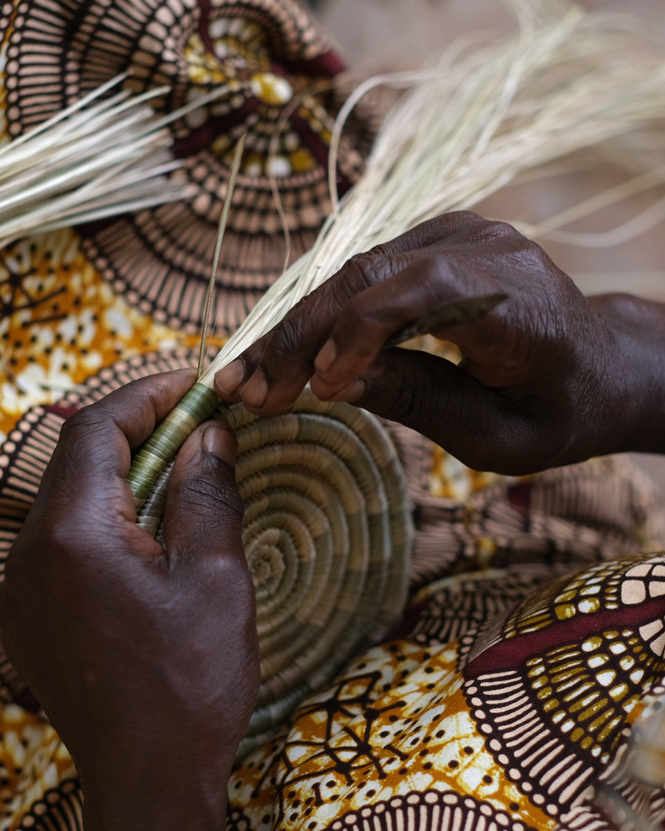 Close-up of a person's hands weaving a round woven basket or tray from natural fibers, with a patterned textile in the background.
