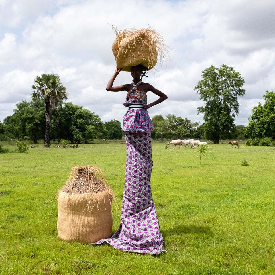 Woman carrying a basket on her head in a grassy field with trees and animals in the background