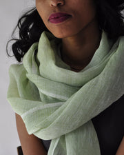 A woman wearing a light sage green linen scarf wrapped around her neck, paired with a black sleeveless top against a plain white wall.