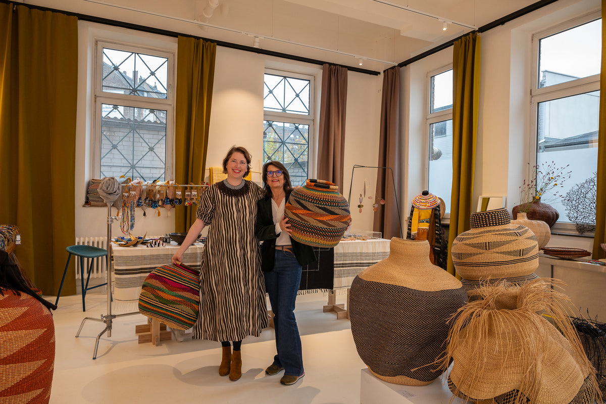 Two women standing in a bright, modern craft studio filled with woven baskets and pottery. The woman on the left wears a long, wide-striped brown and cream dress, while the woman on the right wears a black top and blue jeans.