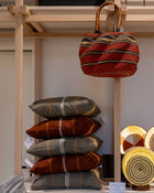 A styled display of woven home goods on a light wood shelving unit, featuring a red and orange patterned hanging tote bag, stacked throw pillows in gray and rust tones, and several round woven coasters or plates with gold-toned bases.