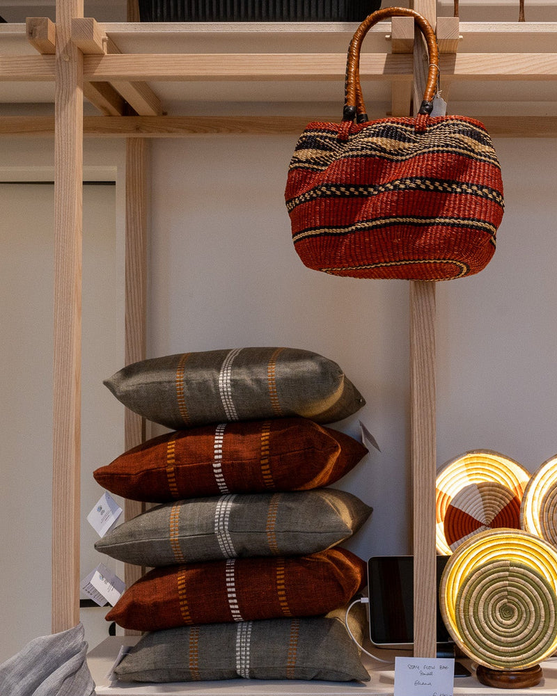 A styled display of woven home goods on a light wood shelving unit, featuring a red and orange patterned hanging tote bag, stacked throw pillows in gray and rust tones, and several round woven coasters or plates with gold-toned bases.