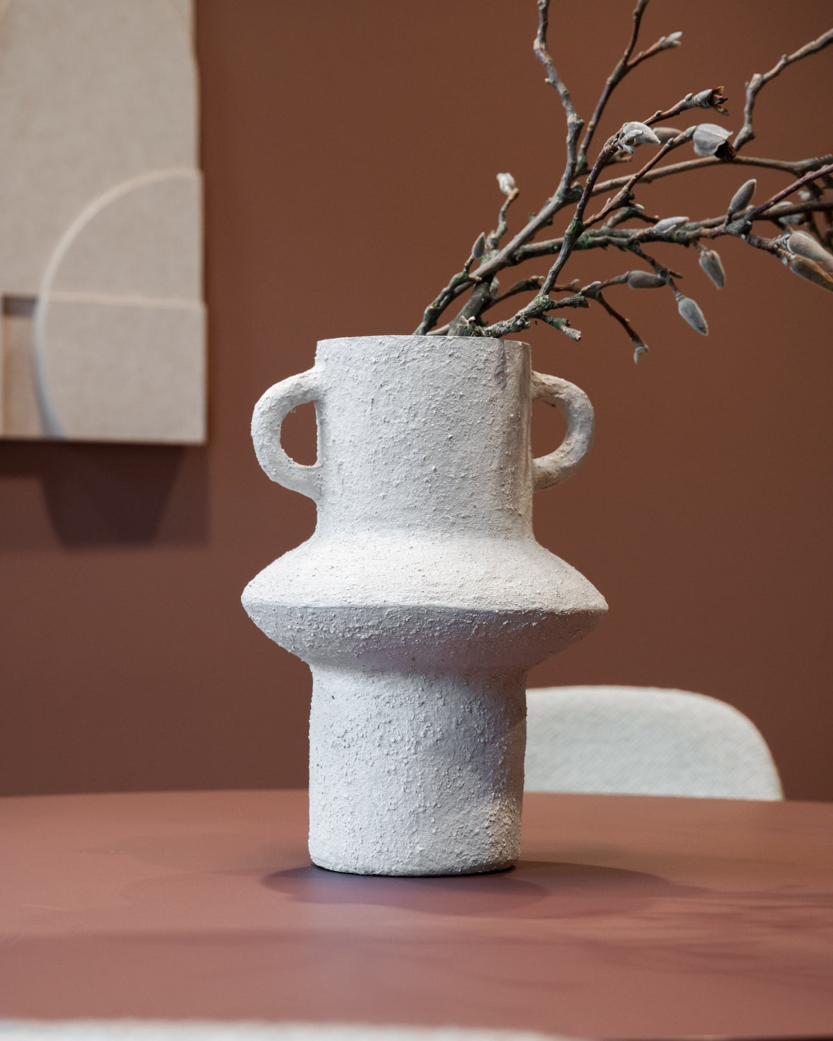 A textured off-white paper pulp vase with two side handles and a bulbous middle section, holding a decorative branch, set on a terracotta-colored table against a warm brown wall.