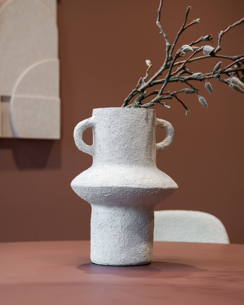 A textured off-white paper pulp vase with two side handles and a bulbous middle section, holding a decorative branch, set on a terracotta-colored table against a warm brown wall.