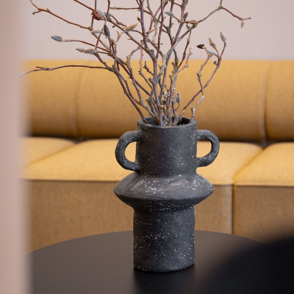 A dark gray textured paper pulp vase with two side handles, filled with dried branches and small buds, placed on a black round side table in front of a yellow sofa.