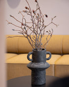 A black textured paper pulp vase with two side handles, filled with dried branches and small buds, placed on a black round side table in front of a yellow sofa.