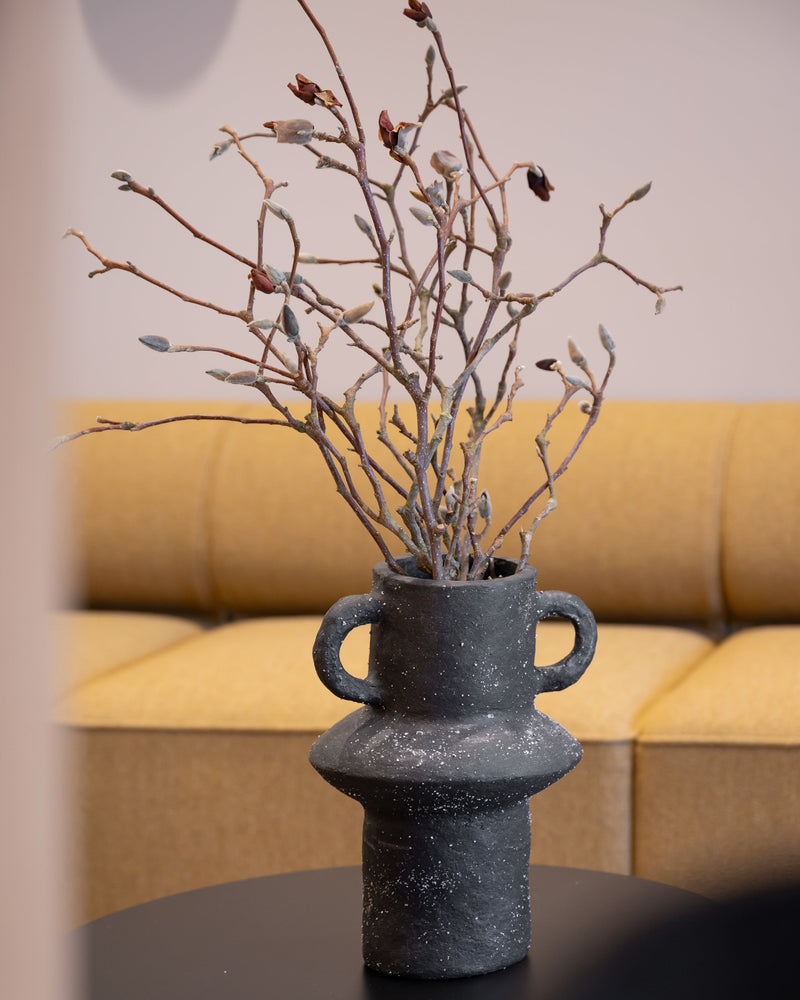 A black textured paper pulp vase with two side handles, filled with dried branches and small buds, placed on a black round side table in front of a yellow sofa.