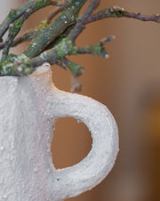 A close-up of a textured, light gray paper pulp planter shaped like a mug with a handle, containing small green-brown tree branches, set against a warm, blurred background.