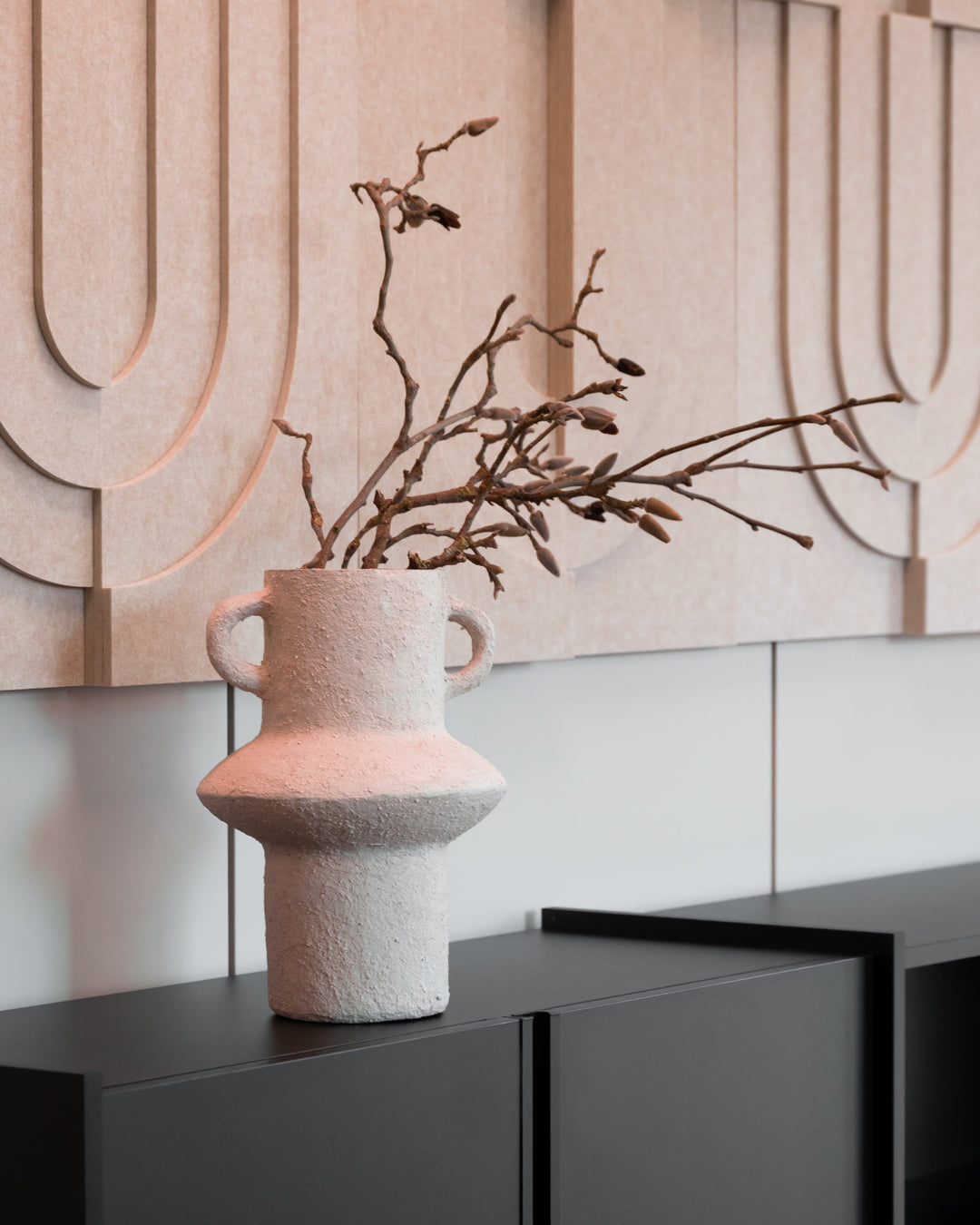 A textured, light pinkish-beige ceramic vase with two side handles and a flared base, holding dried brown branches, displayed on a dark gray cabinet against a modern beige and white wall.