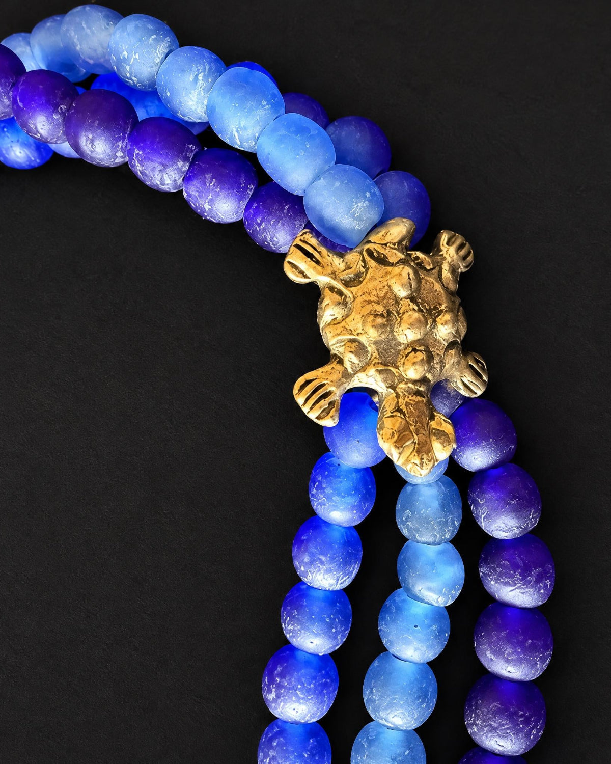 A close-up of a beaded necklace with strands of translucent blue and purple beads, featuring a prominent textured gold turtle charm at the center, set against a solid black background.