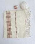 An off-white woven hand towel with blush-colored vertical striped patterns and fringed edges, styled flat-lay on a white textured background with two small decorative seashells.