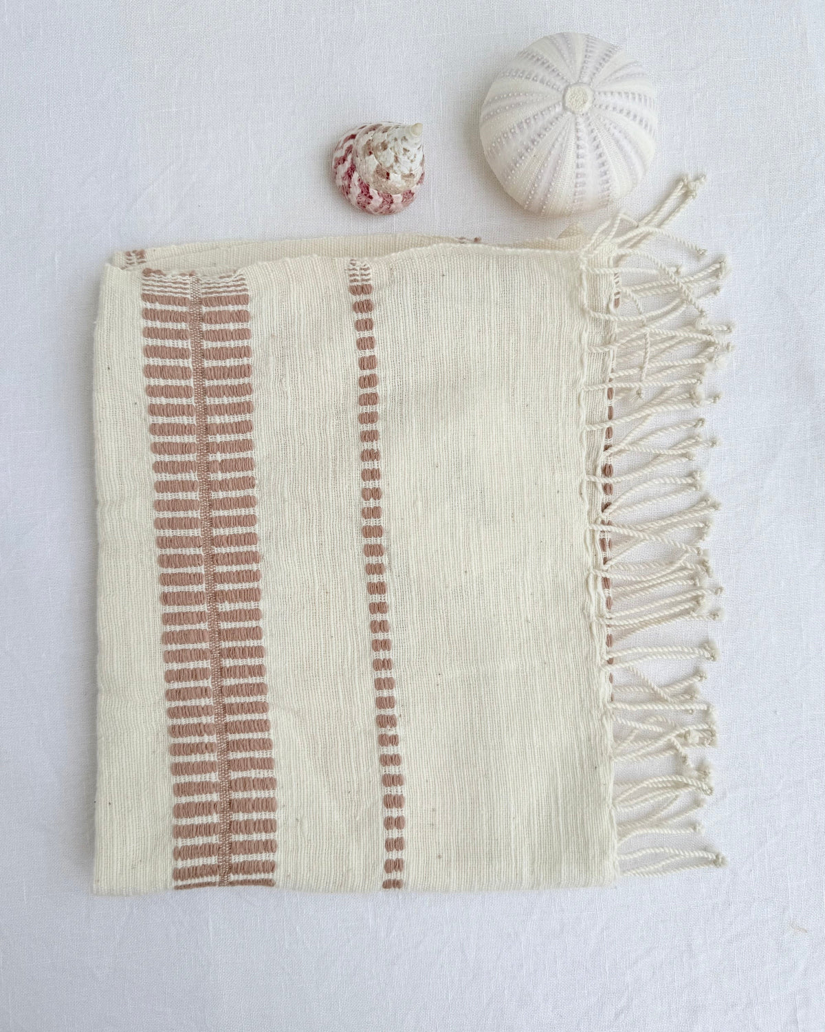 An off-white woven hand towel with blush-colored vertical striped patterns and fringed edges, styled flat-lay on a white textured background with two small decorative seashells.