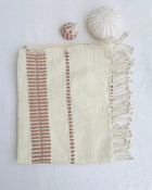 An off-white woven hand towel with blush-colored vertical striped patterns and fringed edges, styled flat-lay on a white textured background with two small decorative seashells.