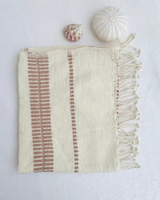 An off-white woven hand towel with blush-colored vertical striped patterns and fringed edges, styled flat-lay on a white textured background with two small decorative seashells.