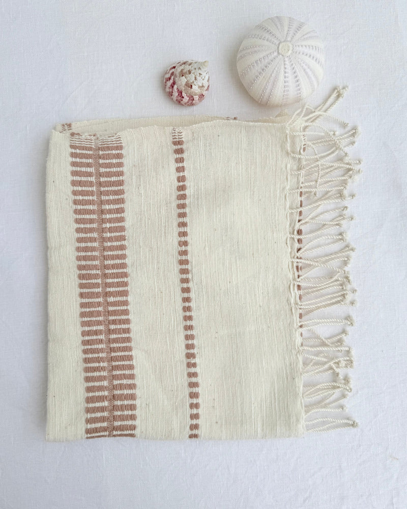 An off-white woven hand towel with blush-colored vertical striped patterns and fringed edges, styled flat-lay on a white textured background with two small decorative seashells.