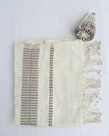 A cream-colored woven hand towel with fringe on one side and vertical gray-striped patterns, styled flat on a white background next to a seashell.