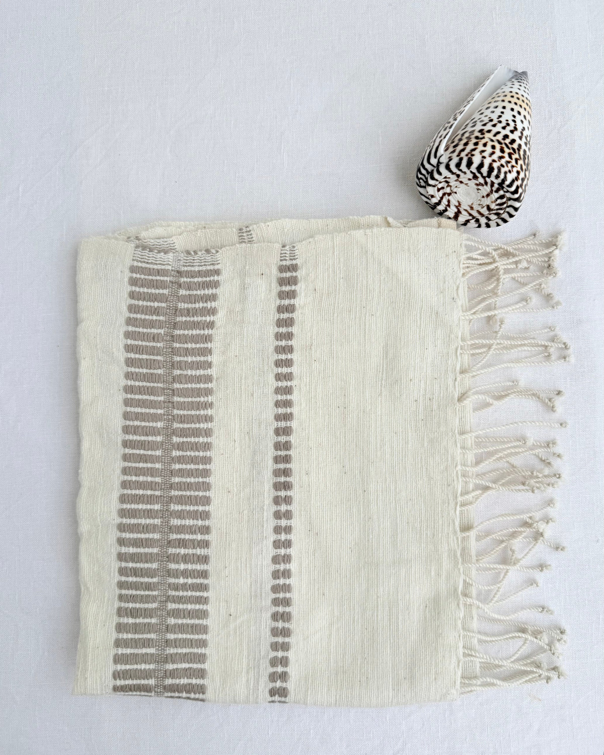 A cream-colored woven hand towel with fringe on one side and vertical gray-striped patterns, styled flat on a white background next to a seashell.