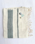 An off-white woven hand towel with fringes, featuring vertical teal-colored stripes. It is styled with three small beach stones on a white textured background.