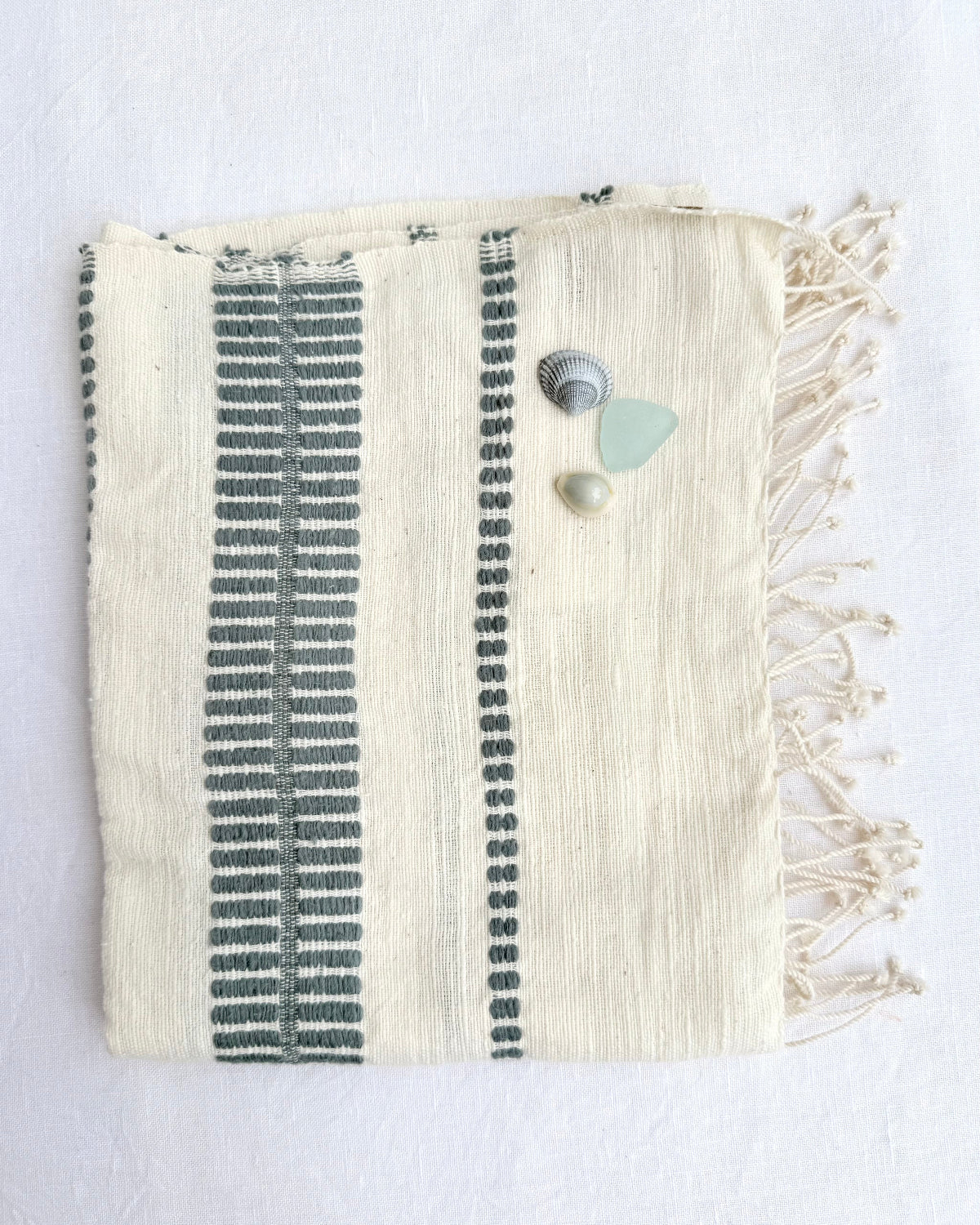 An off-white woven hand towel with fringes, featuring vertical teal-colored stripes. It is styled with three small beach stones on a white textured background.