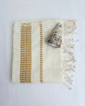 An off-white colored woven hand towel with vertical bronze stripes and fringed edges, styled with a seashell on a white fabric background.
