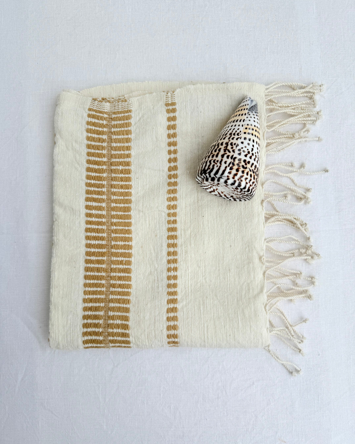 An off-white colored woven hand towel with vertical bronze stripes and fringed edges, styled with a seashell on a white fabric background.