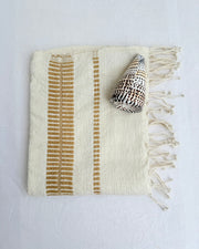 An off-white colored woven hand towel with vertical bronze stripes and fringed edges, styled with a seashell on a white fabric background.