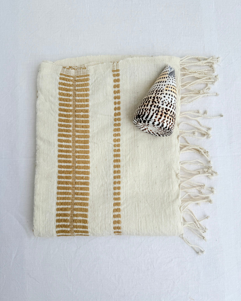 An off-white colored woven hand towel with vertical bronze stripes and fringed edges, styled with a seashell on a white fabric background.