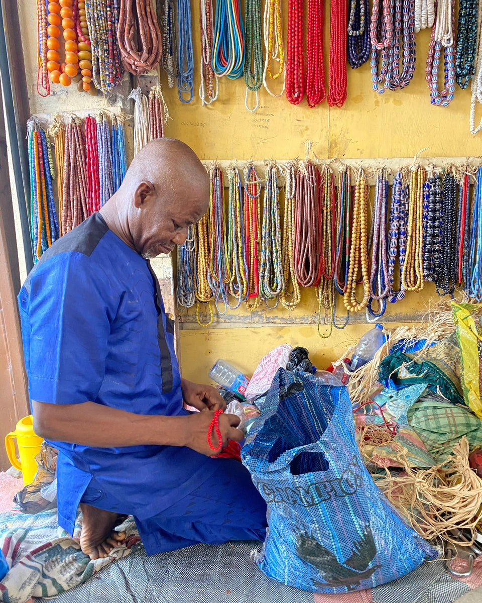 A man in a blue traditional outfit sits on the floor of a bead stall, working with a large blue bag filled with colorful beaded necklaces. The background wall is covered with rows of hanging multicolored beads.