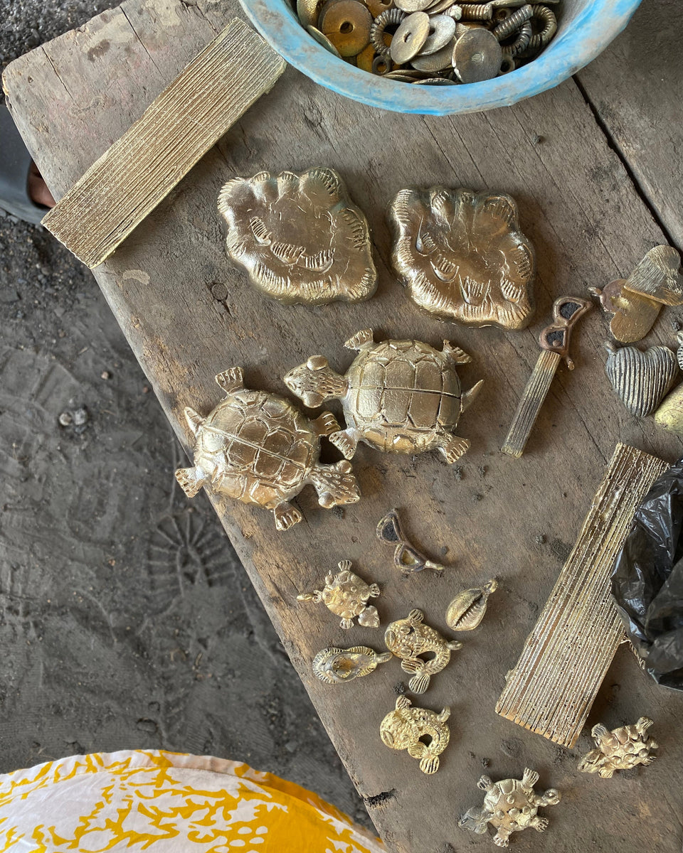 A collection of small brass turtle figurines and decorative hardware pieces arranged on a weathered wooden board, with a blue bowl of coins or disc-like pieces at the top and a patterned yellow-and-white fabric at the bottom left.