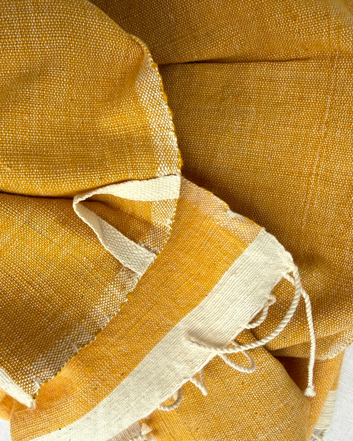 A close-up of a folded mustard yellow woven textile with a contrasting off-white border and frayed fringe, laid on a light neutral background.