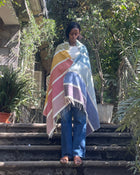 A woman stands on outdoor stone steps surrounded by lush green plants and trees, wrapped in a striped multicolor blanket with white fringe. She is wearing blue jeans and sandals.